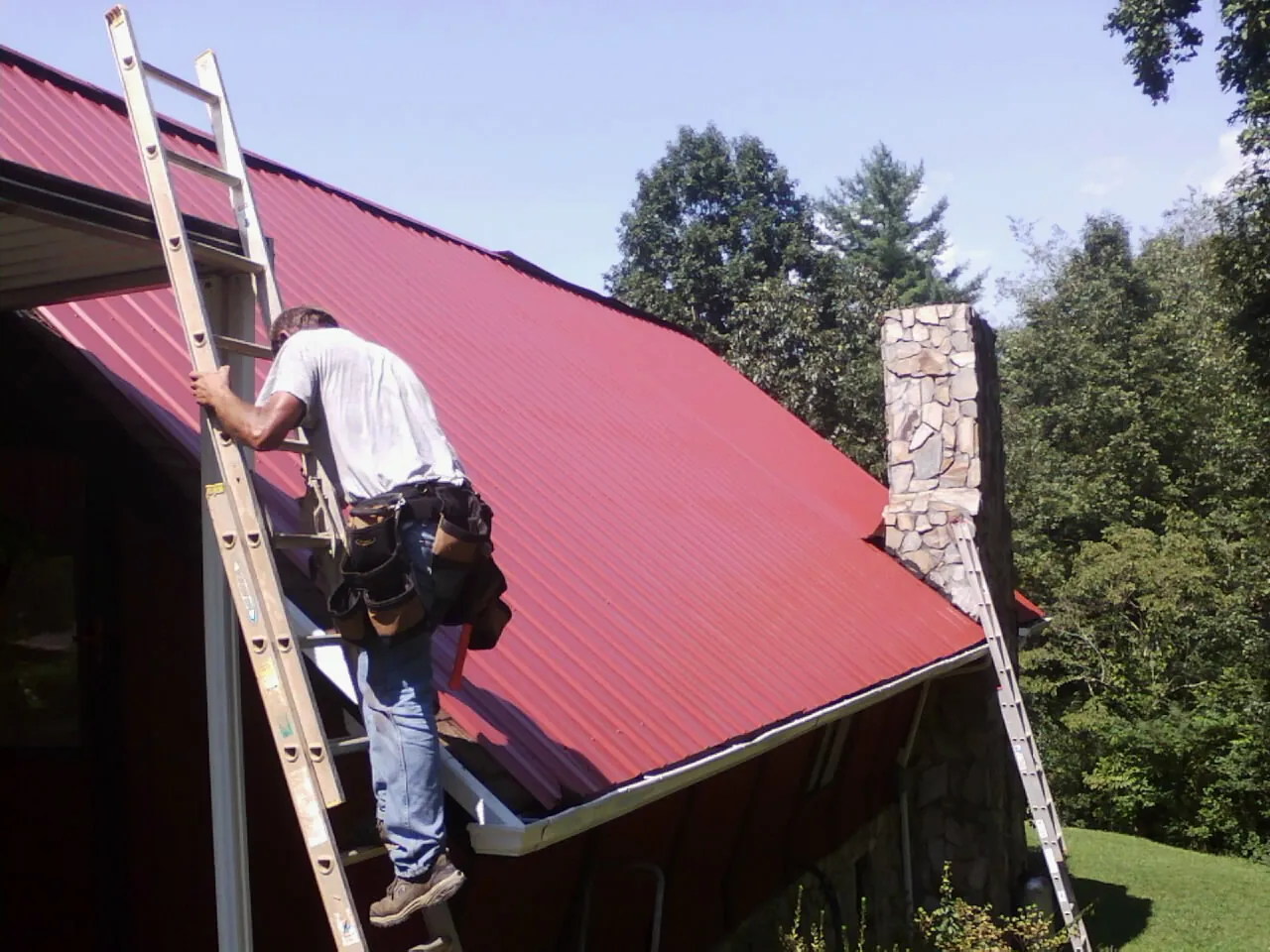 Roofer on a ladder beside a red metal roof and stone chimney — Soffit & Fascia Repair by Roofing Brothers in Midland